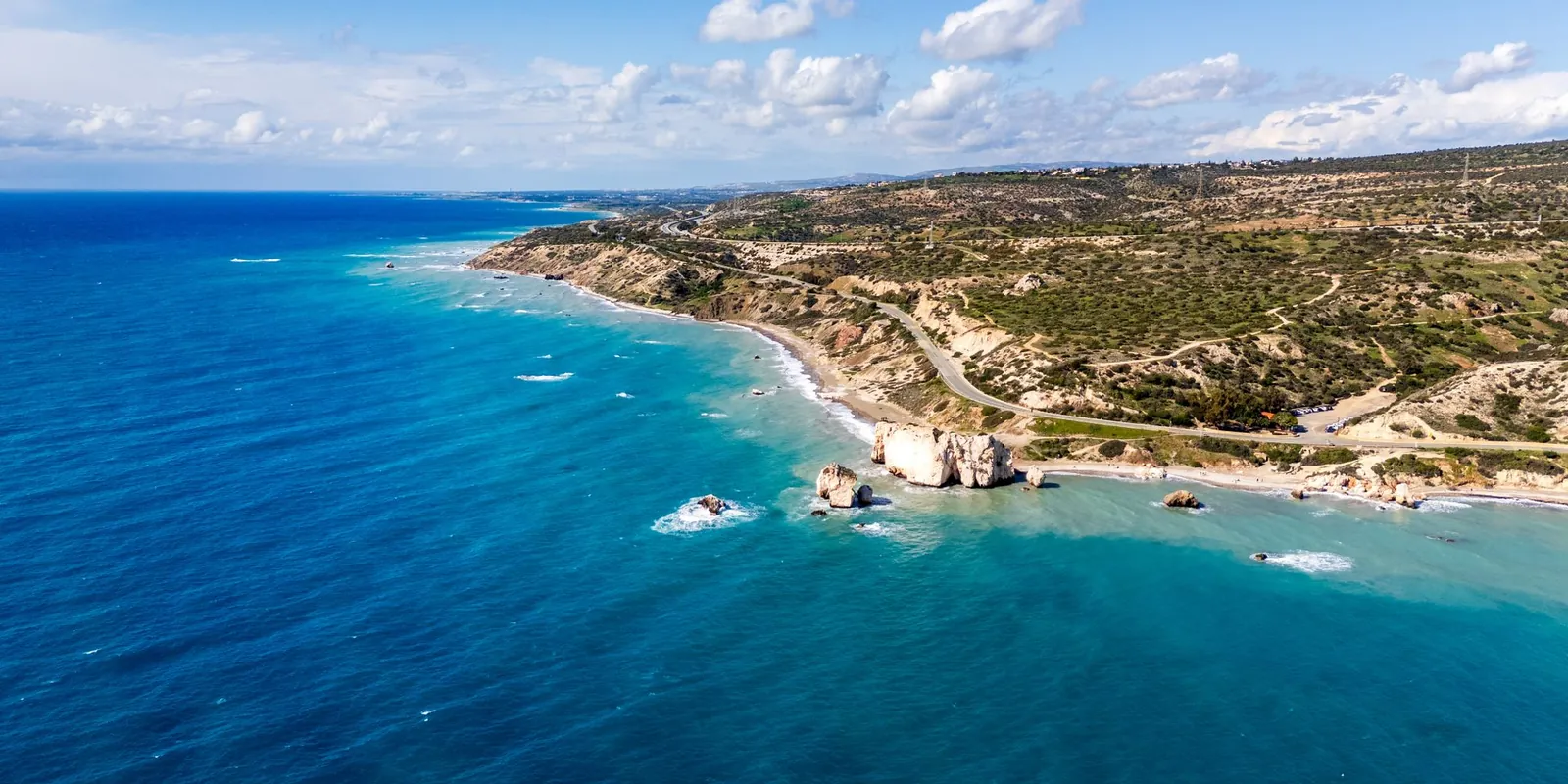 Aerial view of Petra tou Romiou, Aphrodite's Rock, and the green Paphos coastline above a deep blue sea