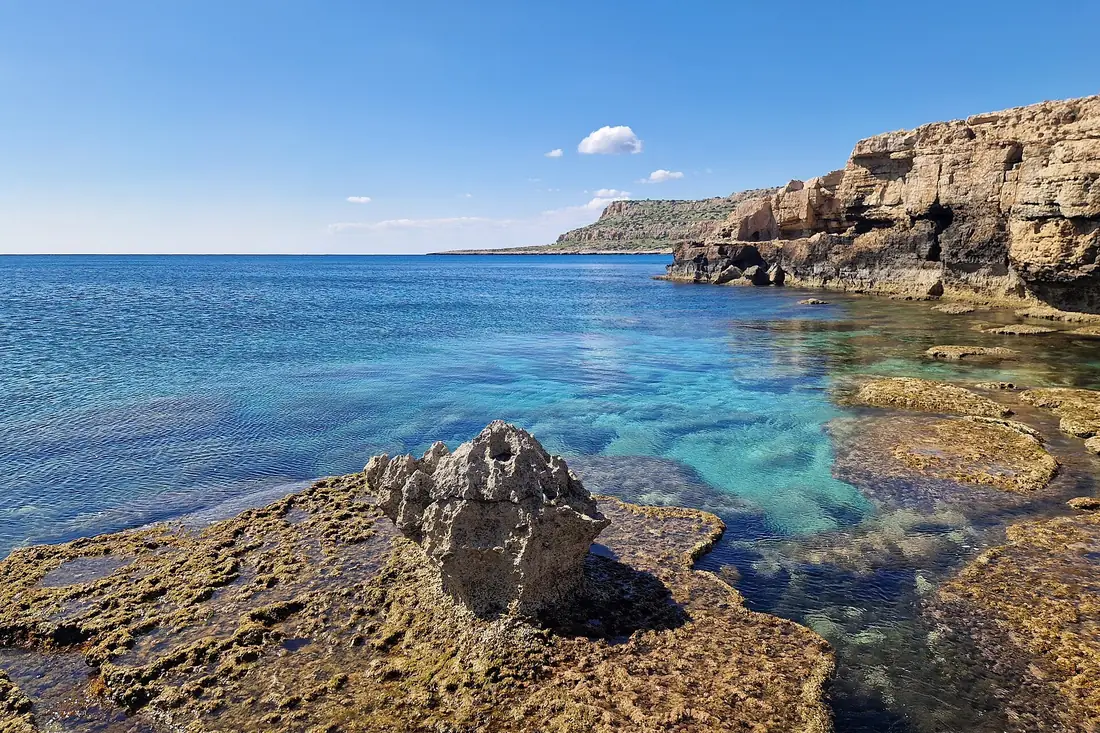 Clear water over flat rocks on the southern shore of Cape Greco, with cliffs in the distance